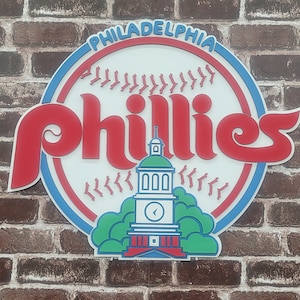 May include: A circular sign displaying the Philadelphia Phillies logo. The sign has a white backdrop with red and blue detailing. The word "Phillies" is in red, and "Philadelphia" is in blue. Independence Hall is illustrated at the bottom.