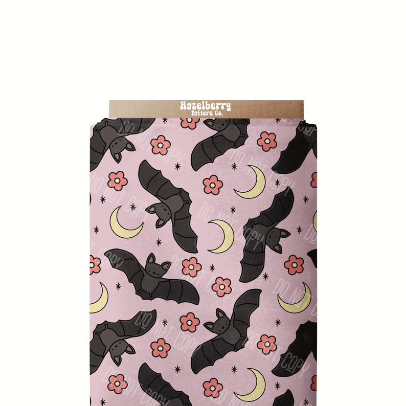 May include: A roll of fabric featuring a repeating pattern of bats, flowers, moons, and stars on a pale pink backdrop. The bats are dark grey, the flowers are pink, and the moons are yellow. The text "Hazelberry Pattern Co." is visible.