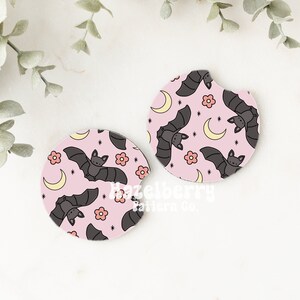 May include: Two pink car coasters featuring a pattern of grey bats, yellow crescent moons, and pink flowers. The coasters are circular with a small notch. The design is whimsical and suitable for Halloween.