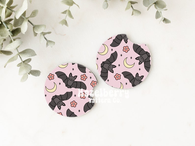 May include: Two pink car coasters featuring a pattern of grey bats, yellow crescent moons, and pink flowers. The coasters are circular with a small notch. The design is whimsical and suitable for Halloween.