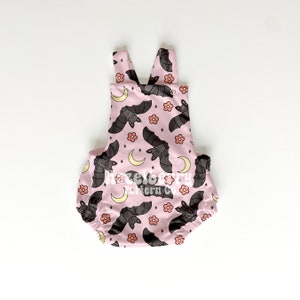May include: Pale pink baby romper featuring a pattern of grey bats, peach flowers, and yellow crescent moons. The romper has a scoop neck and shoulder straps. The text "hazelberry pattern co." is printed on the front.