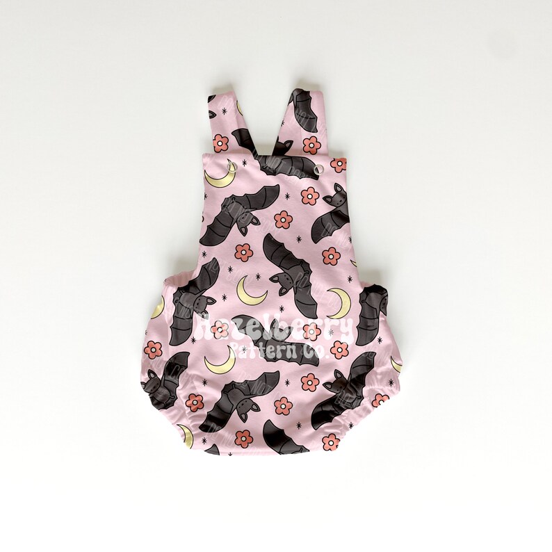 May include: Pale pink baby romper featuring a pattern of grey bats, peach flowers, and yellow crescent moons. The romper has a scoop neck and shoulder straps. The text "hazelberry pattern co." is printed on the front.