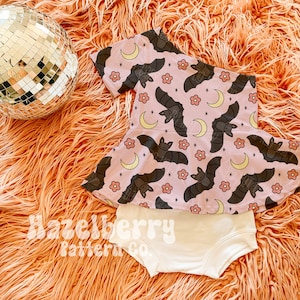 May include: A baby's two-piece outfit. The top is pink with a pattern of bats and moons, and the bottoms are cream-coloured. The outfit is displayed on a fluffy orange surface, with a disco ball. The text "Hazelberry Pattern Co." is visible.