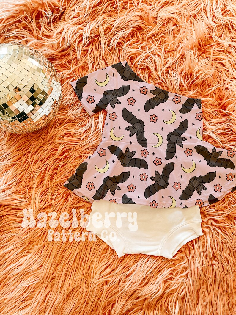 May include: A baby's two-piece outfit. The top is pink with a pattern of bats and moons, and the bottoms are cream-coloured. The outfit is displayed on a fluffy orange surface, with a disco ball. The text "Hazelberry Pattern Co." is visible.