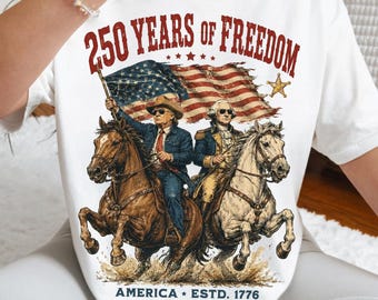 May include: A white short-sleeve t-shirt featuring a graphic print with the text 250 YEARS OF FREEDOM and AMERICA ESTD. 1776. The design depicts two figures on horseback in front of an American flag, with a star icon and a dusty ground element.