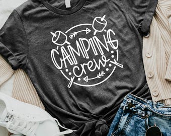 May include: A dark gray short-sleeve t-shirt featuring a white circular graphic with the text "CAMPING crew" and illustrations of marshmallows on sticks. The shirt is displayed alongside a beige cardigan, blue denim shorts, white sneakers, and a black bag.