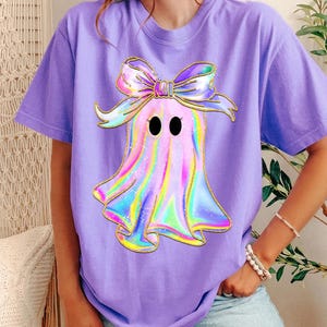 May include: A lavender short-sleeve t-shirt featuring a central graphic of a ghost with a holographic, iridescent finish. The ghost has two black oval eyes and is wearing a large, matching holographic bow on its head with gold outlines.