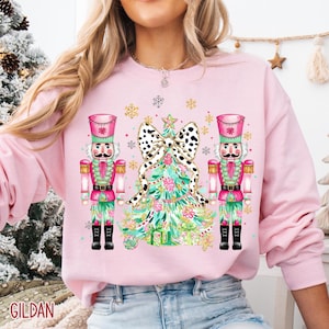 May include: Pink Gildan sweatshirt featuring a Christmas design. The design includes two nutcrackers, a Christmas tree with a dalmatian print bow, and snowflakes. The nutcrackers are pink, green, and red. The Christmas tree is green and pink.