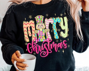 Può includere: Felpa nera con un design colorato "Buon Natale". Il design presenta uno schiaccianoci e testo in rosa, giallo, blu e verde. La felpa è indossata con jeans blu e una tazza bianca.