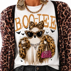 May include: A white t-shirt featuring a graphic of a ghost wearing leopard print sunglasses, a matching bow, a faux fur stole, and a necklace. The ghost holds a coffee cup and a pink leopard print handbag. The text BOO-JEE is printed in orange above.