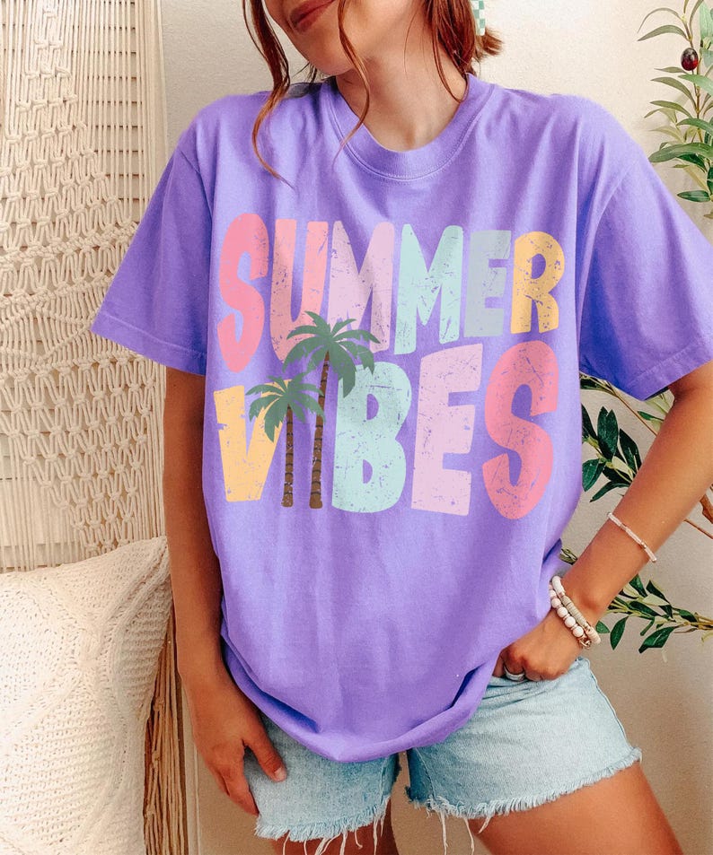 May include: A lilac t-shirt featuring the words "SUMMER VIBES" in pastel shades and palm tree illustrations. The short-sleeved top is made of a soft fabric.