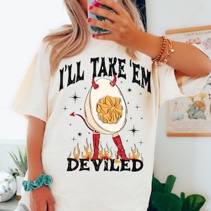 May include: Cream-colored t-shirt with a graphic of a deviled egg wearing red boots and devil horns. The egg has a yolk center and is surrounded by flames. The text 'I'll Take 'Em' is above the egg, and 'Deviled' is below.