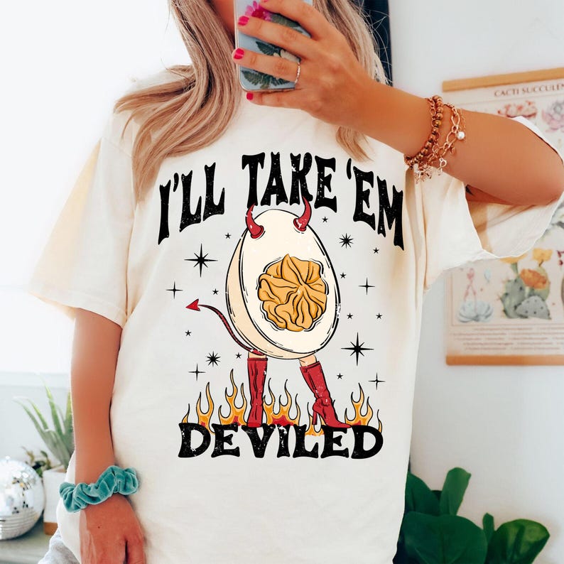 May include: Cream-colored t-shirt with a graphic of a deviled egg wearing red boots and devil horns. The egg has a yolk center and is surrounded by flames. The text 'I'll Take 'Em' is above the egg, and 'Deviled' is below.