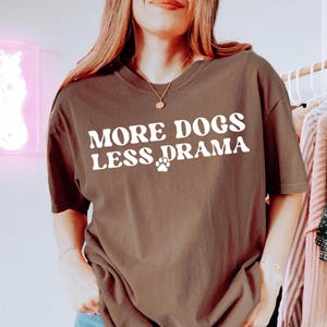 K&ouml;nnte beinhalten: Ein braunes T-Shirt mit dem wei&szlig;en Aufdruck "MORE DOGS LESS DRAMA" und einem Pfotenabdruck. Das l&auml;ssige T-Shirt hat eine lockere Passform und ist wahrscheinlich aus einem weichen Stoff gefertigt. Das Shirt ist Teil einer Bekleidungskollektion.