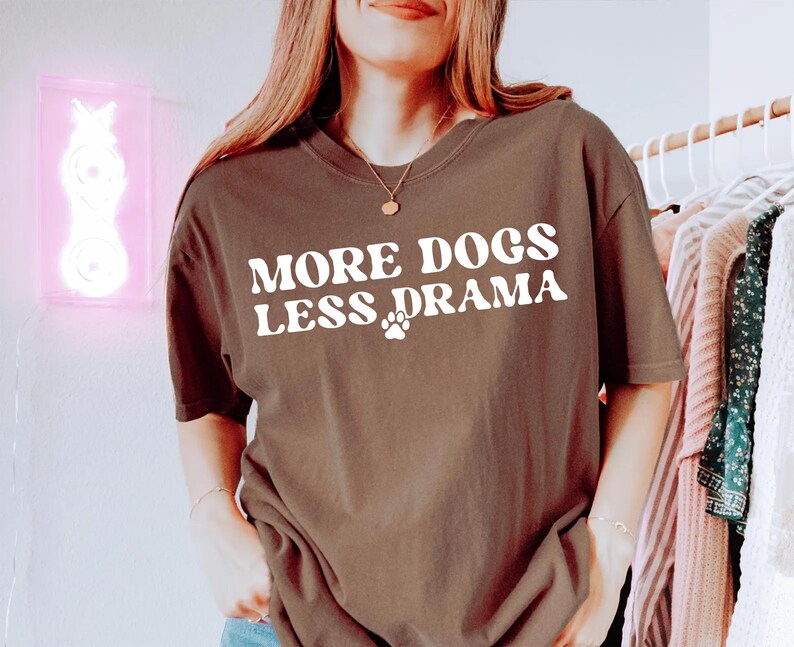 K&ouml;nnte beinhalten: Ein braunes T-Shirt mit dem wei&szlig;en Aufdruck "MORE DOGS LESS DRAMA" und einem Pfotenabdruck. Das l&auml;ssige T-Shirt hat eine lockere Passform und ist wahrscheinlich aus einem weichen Stoff gefertigt. Das Shirt ist Teil einer Bekleidungskollektion.