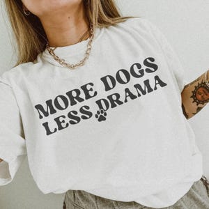 K&ouml;nnte beinhalten: Wei&szlig;es T-Shirt mit dem Text "MORE DOGS LESS DRAMA" in fetter, schwarzer Schrift. Zwischen den W&ouml;rtern befindet sich ein Pfotenabdruck. Das Shirt ist ein l&auml;ssiges Kleidungsst&uuml;ck f&uuml;r den Alltag.