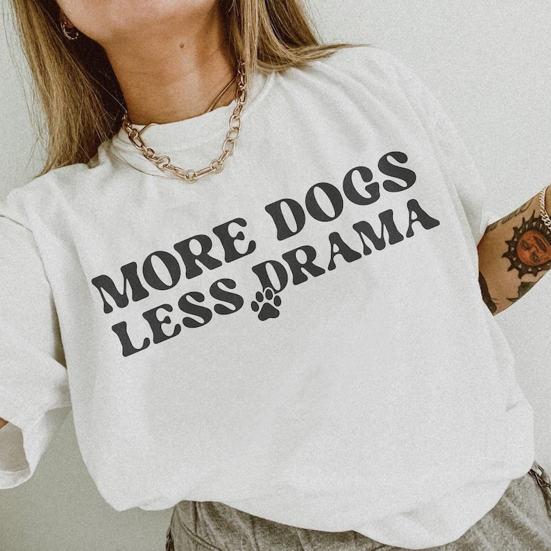 K&ouml;nnte beinhalten: Wei&szlig;es T-Shirt mit dem Text "MORE DOGS LESS DRAMA" in fetter, schwarzer Schrift. Zwischen den W&ouml;rtern befindet sich ein Pfotenabdruck. Das Shirt ist ein l&auml;ssiges Kleidungsst&uuml;ck f&uuml;r den Alltag.