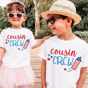 May include: Two children wearing white t-shirts with the text "cousin crew" and a red, white, and blue firework graphic. The girl is wearing a pink tutu skirt and sunglasses. The boy is wearing a straw hat and sunglasses.