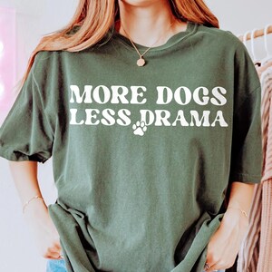 K&ouml;nnte beinhalten: Dunkelgr&uuml;nes T-Shirt mit dem wei&szlig;en Schriftzug &bdquo;MORE DOGS LESS DRAMA&ldquo; und einem wei&szlig;en Pfotenabdruck. Das Shirt hat einen Rundhalsausschnitt und kurze &Auml;rmel. Der Stoff wirkt weich und bequem.