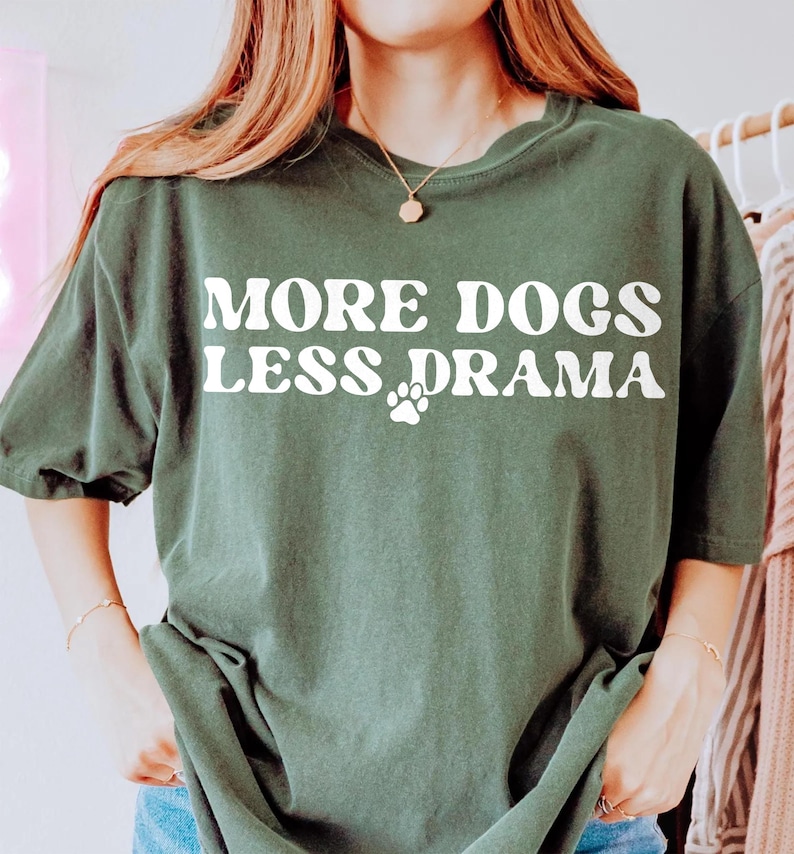 K&ouml;nnte beinhalten: Dunkelgr&uuml;nes T-Shirt mit dem wei&szlig;en Schriftzug &bdquo;MORE DOGS LESS DRAMA&ldquo; und einem wei&szlig;en Pfotenabdruck. Das Shirt hat einen Rundhalsausschnitt und kurze &Auml;rmel. Der Stoff wirkt weich und bequem.