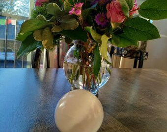 Puede incluir: Una bola de cristal blanca se encuentra sobre una mesa de madera frente a un jarrón lleno de flores rosas y moradas.