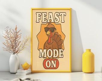 May include: A framed print with a yellow border features a cartoon turkey wearing sunglasses. The text "FEAST MODE ON" is displayed in a retro font. The background is a light orange color. A white vase with dried flowers and a yellow vase are also in the image.