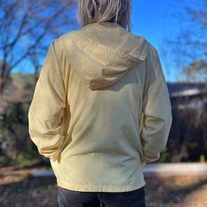 May include: A pale yellow hooded jacket with a zipper closure. The jacket is made of a lightweight, water-resistant material.