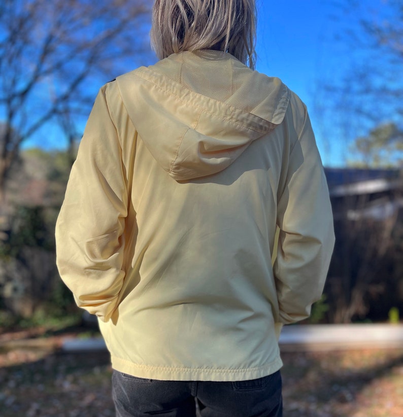 May include: A pale yellow hooded jacket with a zipper closure. The jacket is made of a lightweight, water-resistant material.