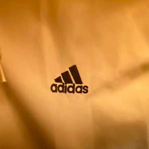May include: A close-up of a cream-colored hoodie with a black Adidas logo embroidered on the front.