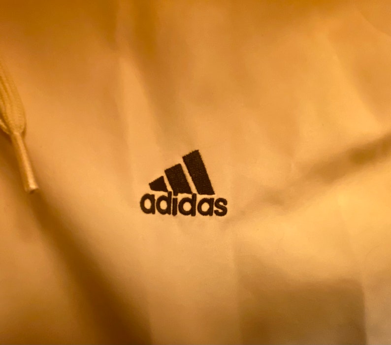 May include: A close-up of a cream-colored hoodie with a black Adidas logo embroidered on the front.