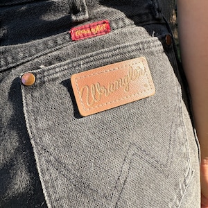 May include: A pair of black denim jeans with a brown leather Wrangler label on the back pocket. The label has the word "Wrangler" written in gold lettering.