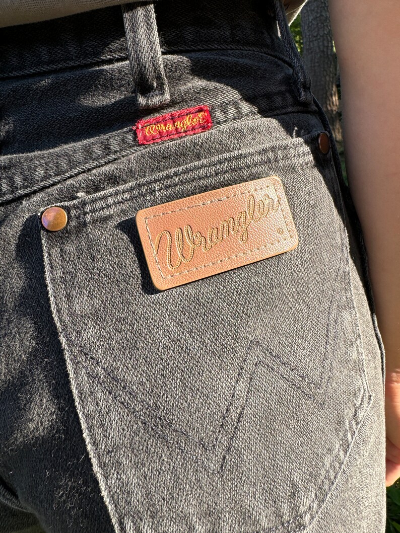 May include: A pair of black denim jeans with a brown leather Wrangler label on the back pocket. The label has the word "Wrangler" written in gold lettering.