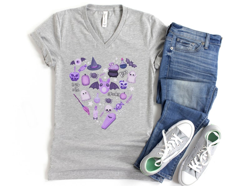 Pu&ograve; includere: Una maglietta grigia melange a collo a V con un design a forma di cuore viola e bianco che presenta varie icone di Halloween, tra cui pipistrelli, fantasmi, zucche, un cappello da strega, un calderone e un teschio. Il testo "Trick or Treat" &egrave; incluso nel design.