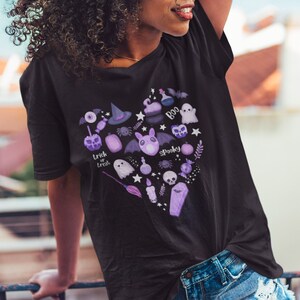 Pu&ograve; includere: Una maglietta nera con un design a forma di cuore viola che presenta varie illustrazioni a tema Halloween, tra cui un fantasma, un pipistrello, un cappello da strega, una zucca, uno scheletro e le parole "trick or treat" e "spooky".