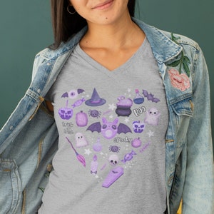 Pu&ograve; includere: Una t-shirt grigia melange a collo a V con un design a forma di cuore viola e bianco che presenta varie icone a tema Halloween, tra cui un cappello da strega, un fantasma, un pipistrello, un ragno, un calderone, una zucca e le parole "Trick or Treat" e "Spooky".