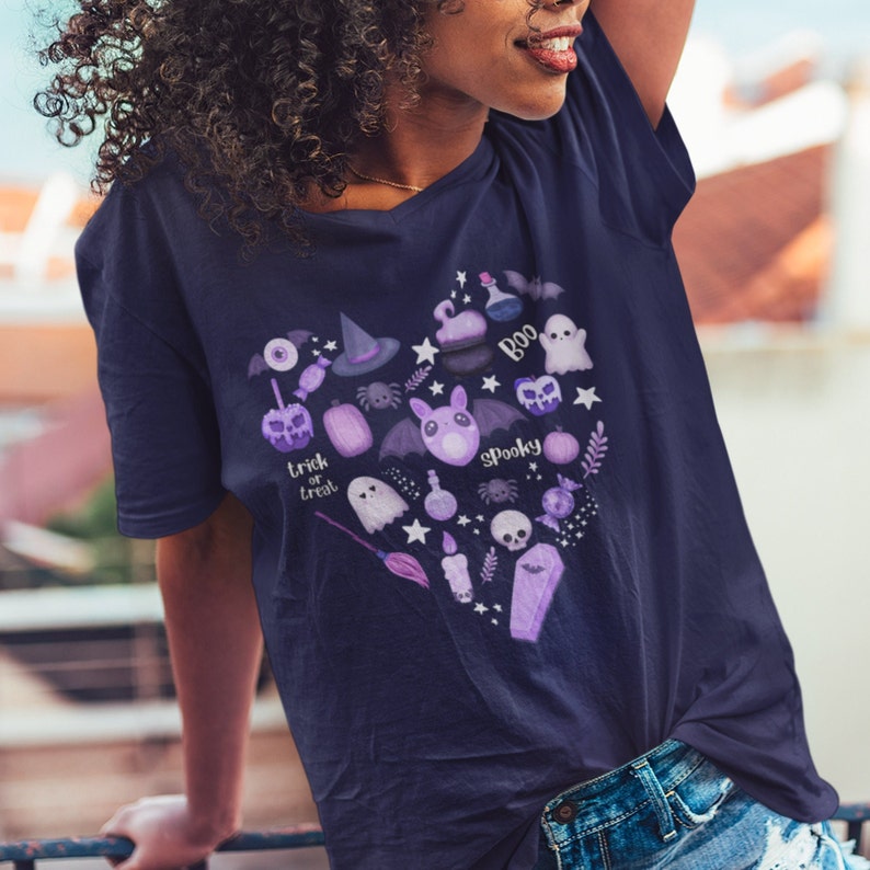 Pu&ograve; includere: Una maglietta blu navy con un design a forma di cuore viola che presenta varie illustrazioni a tema Halloween, tra cui fantasmi, pipistrelli, zucche, un cappello da strega, un ragno, uno scheletro, una bottiglia di pozione, una bara, una candela, una scopa e le parole "trick or treat" e "spooky".