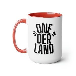May include: White ceramic mug with a red rim and handle. The mug has black text that reads "ONE DER LAND" with stylized water droplets around the word "DER".