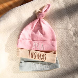 May include: Three personalised baby hats stacked on top of each other. The top hat is pink with the name "Ayla" embroidered in white. The middle hat is beige with the name "Thomas" embroidered in brown. The bottom hat is light blue with the name "Evelyn" embroidered in white.