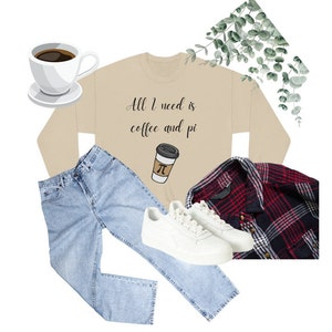 May include: A beige sweatshirt with the text "All I need is coffee and pi" and a coffee cup graphic. The image also includes light blue jeans, white trainers, a red and black plaid shirt, and a cup of coffee.