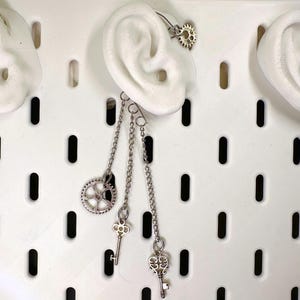 May include: A silver-toned steampunk ear cuff featuring a gear design that curves over the ear. Dangling from the cuff are three chains with a gear and two key charms. The ear cuff is displayed on a white ear model.