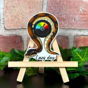 May include: A sticker featuring a cartoon snake curled up with a rainbow-colored dial that reads "Lazy Day". The sticker is on a small wooden easel.