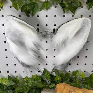 May include: A pair of white and gray faux fur animal ears attached to a metal headband. The ears are fluffy and have a black outline. The background is a white pegboard with green leaves.