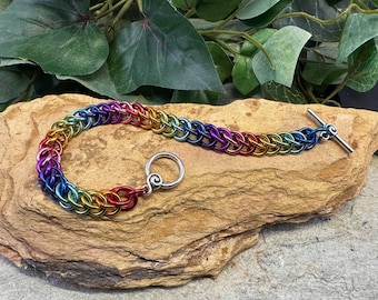May include: A rainbow-colored chainmail bracelet with a toggle clasp. The bracelet is made of small, interlocking metal rings in a spectrum of colors, including red, orange, yellow, green, blue, and purple. The bracelet is displayed on a textured, light brown stone.