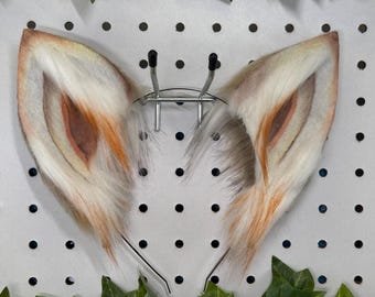 May include: A pair of fox ears with a realistic design. The ears are primarily white with brown and orange accents, and are attached to a metal headband. The ears are displayed against a white background with black dots and green leaves.