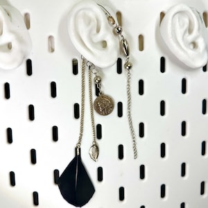 May include: An ear cuff with a dangling chain, coin pendant, and black feather. The ear cuff features a series of beads and a chain that drapes down. The coin pendant has a profile image. The black feather adds a dramatic flair.