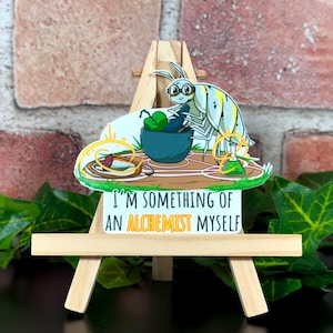May include: A sticker depicting a cartoon insect with glasses, a mortar and pestle, and a beaker. The text "I'M SOMETHING OF AN ALCHEMIST MYSELF" is displayed below the illustration. The sticker is mounted on a small wooden easel.