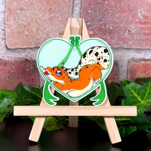 May include: A sticker featuring a cartoon gecko inside a heart-shaped frame. The gecko is orange with black spots and a pink tongue. The heart is light blue with a green border and ribbon design.