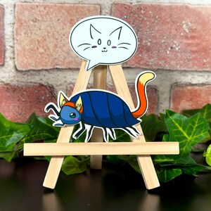 May include: A whimsical sticker set featuring a blue and orange bug-like creature with a fox tail, and a white speech bubble with a cat face. The stickers are displayed on a small wooden easel, with a brick background and green foliage.