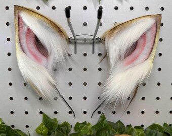 May include: A pair of faux fur animal ears, featuring a tan and white color scheme with pink inner ear detailing. The ears are mounted on a metal headband, designed to be worn. The ears are fluffy and detailed.