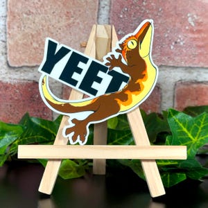 May include: A sticker depicting a cartoon gecko with brown, yellow, and orange markings, holding a sign that reads "YEET" in bold, black letters. The gecko is displayed on a small wooden easel, with a brick background and green foliage.